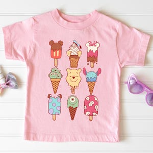 May include: Pink short-sleeved t-shirt featuring cartoon ice cream popsicles in various designs. The popsicles include Mickey Mouse, Winnie the Pooh, and other characters. The shirt is made of a soft material.