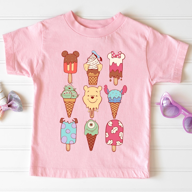 May include: Pink short-sleeved t-shirt featuring cartoon ice cream popsicles in various designs. The popsicles include Mickey Mouse, Winnie the Pooh, and other characters. The shirt is made of a soft material.