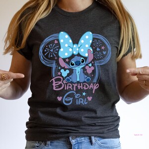 May include: Dark gray t-shirt featuring a blue cartoon character with a polka dot bow, surrounded by a Mickey Mouse-shaped design. The text "Birthday Girl" is in pink and blue. The shirt is a casual wear item.