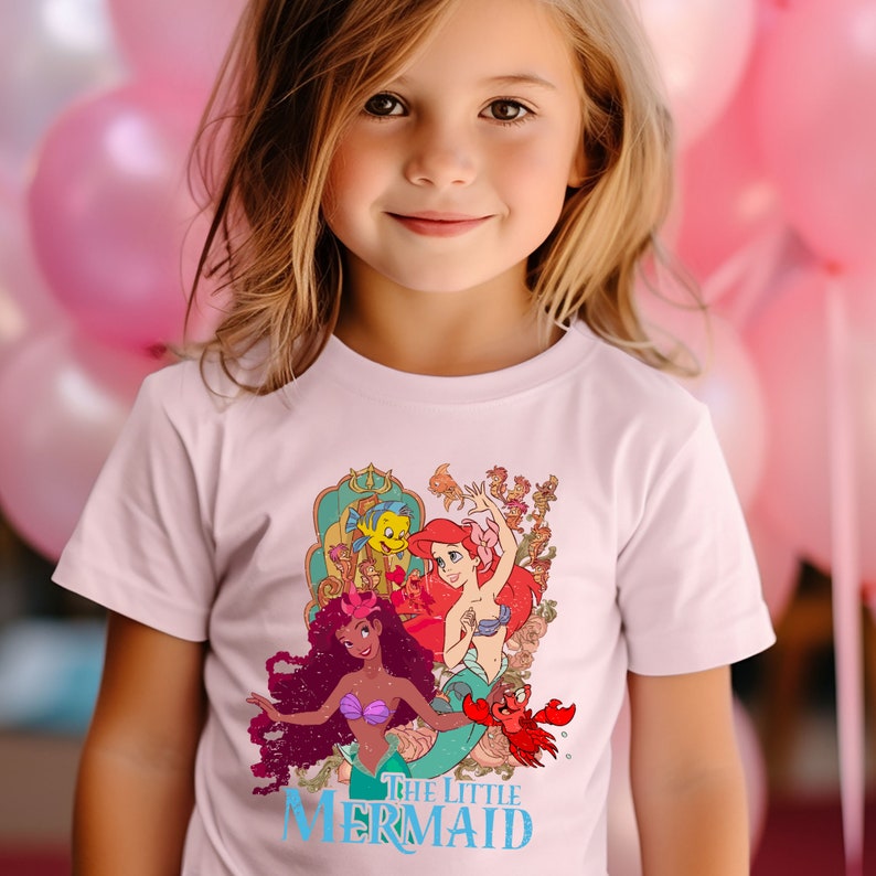 Puede incluir: Una camiseta blanca con un gr&aacute;fico colorido de Ariel, la Sirenita, y otros personajes de la pel&iacute;cula de Disney. El texto "The Little Mermaid" est&aacute; impreso en azul debajo del gr&aacute;fico.
