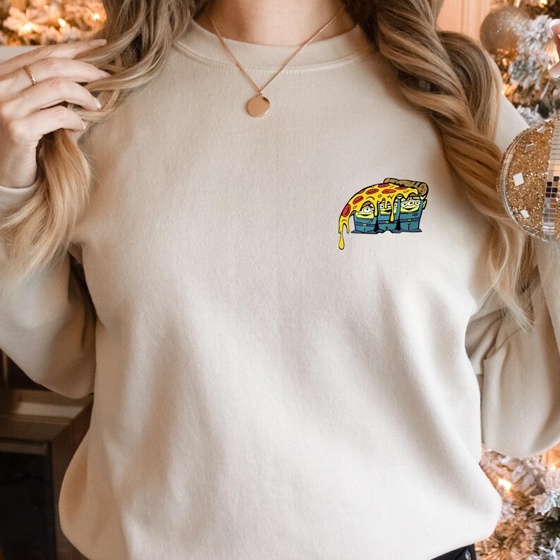 May include: Cream-colored sweatshirt with a small, colorful pizza slice graphic on the chest. The pizza slice has melted cheese and a cartoon face. The sweatshirt is a casual, comfortable style.