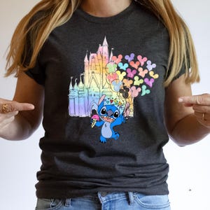 Puede incluir: Camiseta gris oscuro con una colorida ilustraci&oacute;n de Stitch sosteniendo helado y globos frente a un castillo arco&iacute;ris. Los globos tienen forma de cabezas de Mickey Mouse. Un dise&ntilde;o divertido y caprichoso.