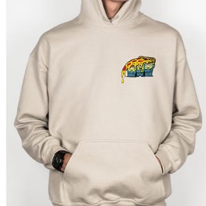 May include: Beige hoodie with a front pocket and a drawstring hood. The hoodie features a colorful graphic of a pizza slice dripping over three cartoon characters. The graphic is on the upper left chest area.