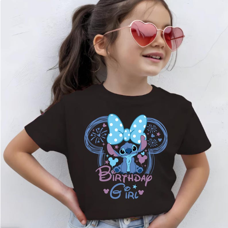 May include: Black t-shirt featuring a cartoon character with a blue bow and the text "Birthday Girl". The design includes blue and pink accents. The shirt is a short-sleeved crew neck.