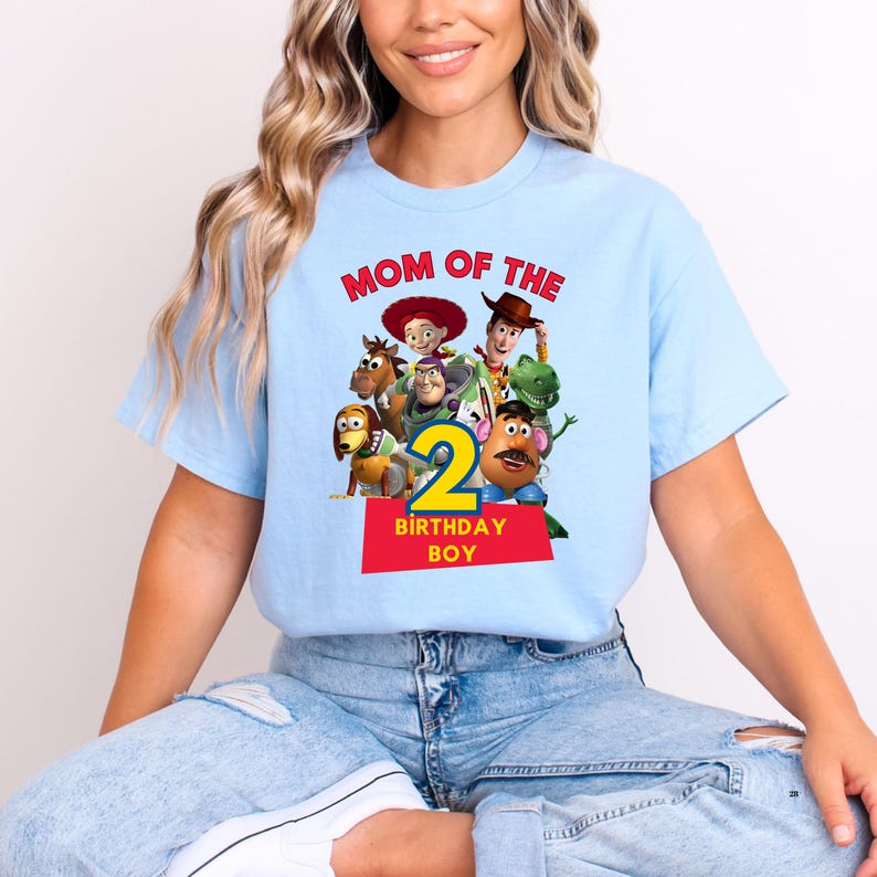 May include: Light blue t-shirt featuring a graphic of characters from a popular animated movie. The shirt has the text "MOM OF THE" above the characters and "2 BIRTHDAY BOY" below. The design is in red, yellow, and green.