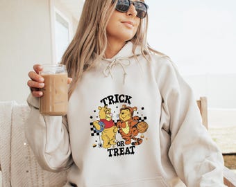 May include: A cream-colored hoodie with a graphic of Winnie the Pooh and Tigger dressed up for Halloween. The graphic says "Trick or Treat" and features a checkered background.