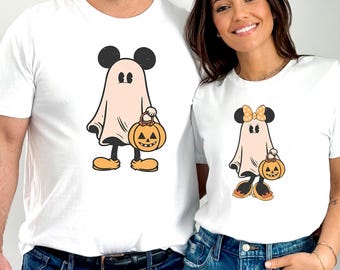 May include: White t-shirts featuring a Halloween-themed design. The design depicts a ghost with Mickey Mouse ears holding a jack-o'-lantern. One shirt has a bow on the ears. The shirts are paired with blue jeans.