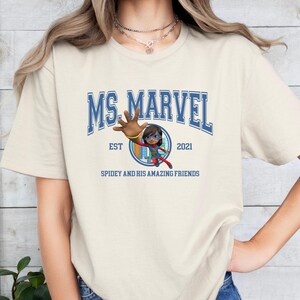 Pu&ograve; includere: T-shirt color crema con la scritta blu "MS. MARVEL" e "EST 2021". Sotto, un'immagine a cartoni animati di Ms. Marvel e la scritta "SPIDEY AND HIS AMAZING FRIENDS". La maglietta ha maniche corte.