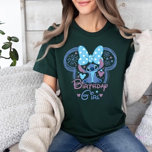 May include: Dark green t-shirt featuring a cartoon character with a blue bow and the text "Birthday Girl". The design includes a blue outline of Mickey Mouse ears and stars. The shirt is made of a soft material.