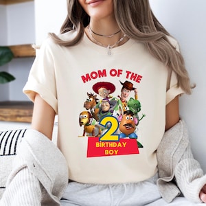 May include: Cream-colored t-shirt featuring a colorful graphic of characters from a popular animated movie. The design includes the text "MOM OF THE" above a number "2" and the words "BIRTHDAY BOY" on a red rectangle.