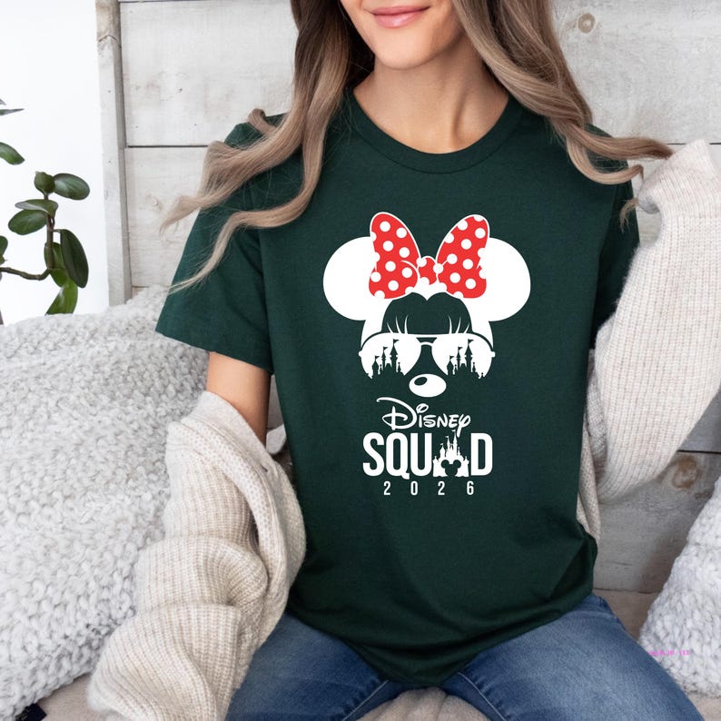 May include: Dark green t-shirt featuring a white Minnie Mouse design with a red polka dot bow, sunglasses, and a silhouette of a castle. The text "Disney Squad 2026" is printed below the graphic. The shirt is a crew neck.