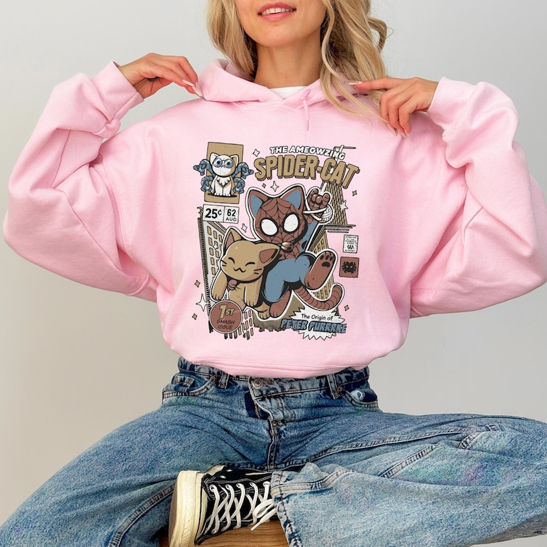 Puede incluir: Sudadera con capucha rosa con un estampado gr&aacute;fico de un gato de dibujos animados superh&eacute;roe con el texto "The Ameowzing Spider-Cat" y "The Origin of Peter Purrke".