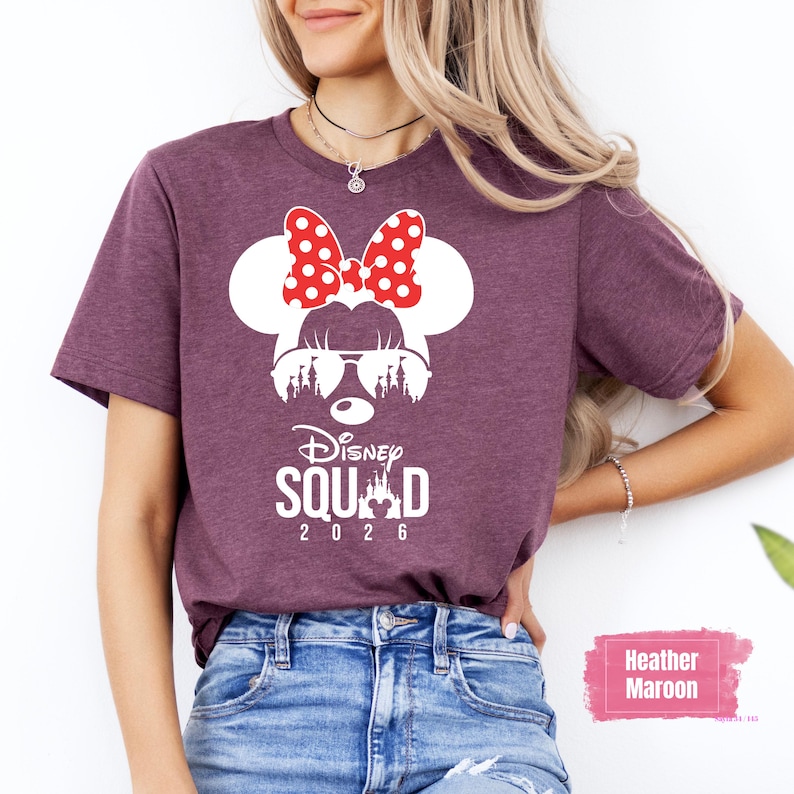 May include: Heather maroon t-shirt featuring a white Minnie Mouse design with a red polka dot bow, sunglasses, and a castle silhouette. The text "Disney Squad 2026" is printed below the graphic. The shirt is worn by a person.