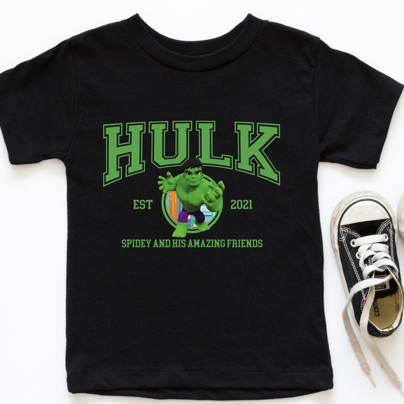 Pu&ograve; includere: T-shirt nera con la scritta "HULK" in lettere verdi. Sotto, il testo "EST 2021" e la frase "SPIDEY AND HIS AMAZING FRIENDS". Un personaggio dei cartoni animati di Hulk &egrave; al centro.