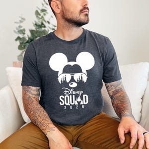 May include: Dark gray t-shirt featuring a white Mickey Mouse silhouette wearing sunglasses with a castle design. The text "Disney Squad 2026" is printed below the graphic. The shirt is made of a soft material.