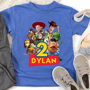 May include: A blue t-shirt featuring a graphic of Toy Story characters, the number 2, and the name DYLAN in white letters on a red rectangle. The shirt is paired with gray pants and gray sneakers.