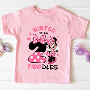 K&ouml;nnte beinhalten: Rosaes Kleinkind-T-Shirt mit Minnie Mouse-Motiv. Der Aufdruck enth&auml;lt den Text "SISTER OF THE 2 TWODLES" in Rosa und Schwarz, eine gepunktete Schleife und die Minnie Mouse-Figur. Das Shirt hat kurze &Auml;rmel und einen Rundhalsausschnitt.