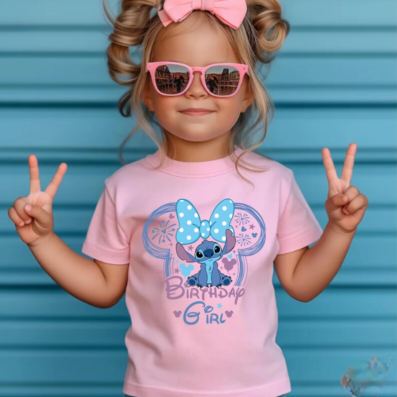 May include: A pink t-shirt featuring a cartoon character with a blue bow and the words "Birthday Girl" in blue script. The shirt is worn by a young child wearing pink sunglasses and a pink bow.