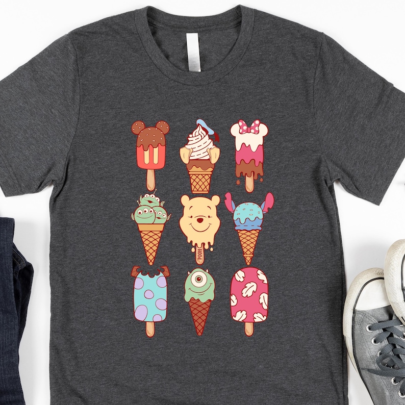 May include: Dark gray t-shirt featuring colorful cartoon ice cream designs. The ice cream treats have various character-inspired designs, including a bear, mouse, and alien. The shirt is made of a soft material and has a classic crew neck.