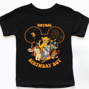 May include: Black t-shirt featuring a colorful graphic of characters from The Lion King, including Simba, Timon, and Pumbaa. The design includes the text "RECHAL", the number "3", and "BIRTHDAY BOY" in a Mickey Mouse-inspired design.