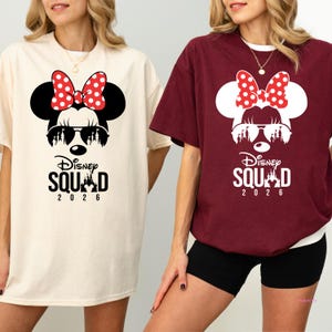 May include: Two t-shirts, one cream and one burgundy, each featuring a Minnie Mouse graphic. The graphic includes a red polka dot bow, sunglasses, and the text "Disney Squad 2026".