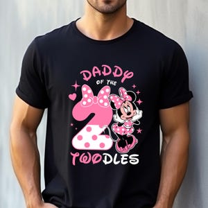 K&ouml;nnte beinhalten: Schwarzes T-Shirt mit einem rosa Aufdruck mit Minnie Mouse, einer gro&szlig;en Zahl 2 und dem Text "DADDY OF THE TWOODLES". Minnie Mouse tr&auml;gt ein rosa gepunktetes Kleid und eine Schleife. Das Design enth&auml;lt Herzen und Sterne.