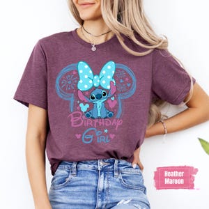 May include: Heather maroon t-shirt featuring a cartoon character with a blue bow and the words "Birthday Girl" in a playful font. The design includes blue accents and hearts. The shirt is paired with blue jeans.