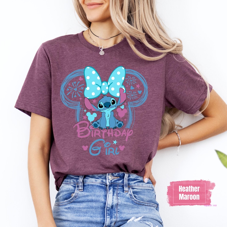 May include: Heather maroon t-shirt featuring a cartoon character with a blue bow and the words "Birthday Girl" in a playful font. The design includes blue accents and hearts. The shirt is paired with blue jeans.