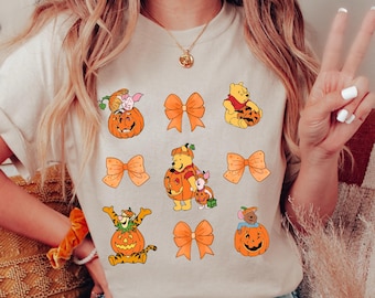 Op de afbeelding: Een licht beige T-shirt met een Winnie de Poeh Halloween-thema. Het shirt toont Winnie de Poeh, Biggetje, Tijger en Eeyore verkleed als pompoenen met oranje strikken.