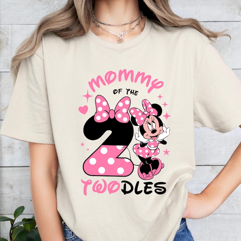 K&ouml;nnte beinhalten: Cremefarbenes T-Shirt mit Minnie Mouse-Grafik und dem Text "Mommy of the 2 Toodles". Die Zahl 2 ist rosa mit wei&szlig;en Punkten. Minnie Mouse tr&auml;gt ein rosa gepunktetes Kleid und eine Schleife.