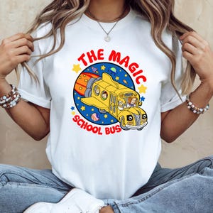 May include: White t-shirt featuring a yellow school bus design with the text "THE MAGIC SCHOOL BUS". The bus is depicted as a spaceship with children inside, surrounded by stars and planets. The design is colorful and cartoonish.