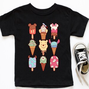 May include: Black short-sleeved t-shirt featuring colorful cartoon ice cream designs. The ice cream treats have various character-inspired shapes and colors, including Mickey Mouse, Winnie the Pooh, and Stitch. A pair of black and white sneakers is visible in the corner.