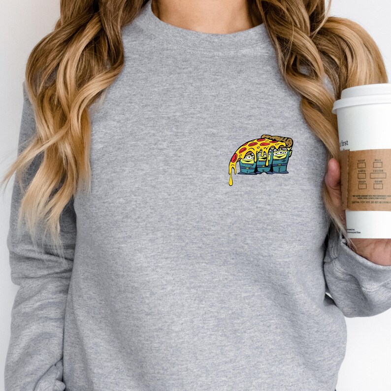 May include: A gray sweatshirt featuring a colorful cartoon pizza slice graphic on the chest. The pizza has melted cheese and is resting on top of three green aliens. The sweatshirt has long sleeves and a crew neck.