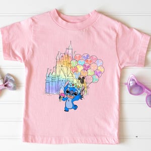 Puede incluir: Una camiseta rosa claro con una ilustraci&oacute;n de dibujos animados de Stitch sosteniendo un helado y globos frente a un castillo colorido. Los globos tienen forma de cabezas de Mickey Mouse.