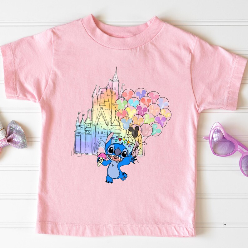 Puede incluir: Una camiseta rosa claro con una ilustraci&oacute;n de dibujos animados de Stitch sosteniendo un helado y globos frente a un castillo colorido. Los globos tienen forma de cabezas de Mickey Mouse.