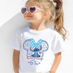 May include: White t-shirt featuring a blue cartoon character with a large blue bow and the words "Birthday Girl." The design is set within a blue outline of mouse ears. The shirt is worn by a child wearing pink heart-shaped sunglasses.