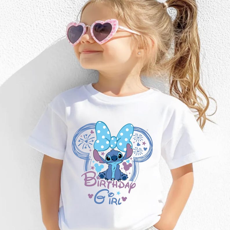 May include: White t-shirt featuring a blue cartoon character with a large blue bow and the words "Birthday Girl." The design is set within a blue outline of mouse ears. The shirt is worn by a child wearing pink heart-shaped sunglasses.