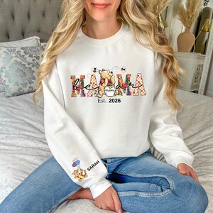 May include: White sweatshirt with the word "Mama" in a colorful floral design, featuring Winnie the Pooh holding a honey pot, with "Est. 2026" below. The sleeve has a cartoon tiger with balloons and the name "Sarah".