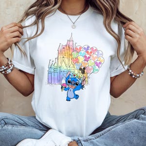 Puede incluir: Camiseta blanca con una ilustraci&oacute;n colorida de Stitch de Lilo & Stitch. Stitch sostiene un helado y globos frente a un castillo arco&iacute;ris. El dise&ntilde;o es caprichoso y juguet&oacute;n, perfecto para los fans de Disney.