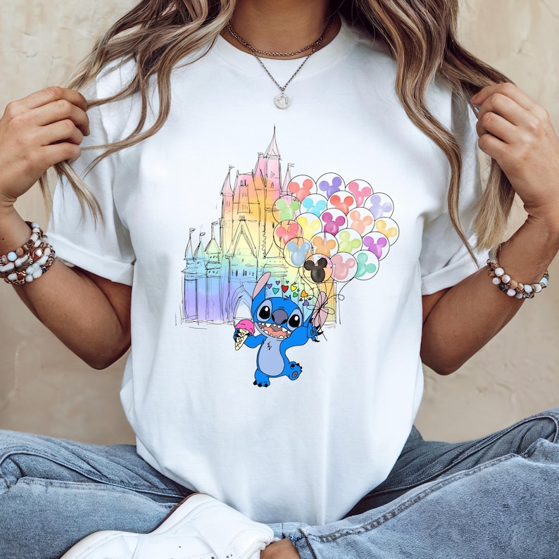 Puede incluir: Camiseta blanca con una ilustraci&oacute;n colorida de Stitch de Lilo & Stitch. Stitch sostiene un helado y globos frente a un castillo arco&iacute;ris. El dise&ntilde;o es caprichoso y juguet&oacute;n, perfecto para los fans de Disney.