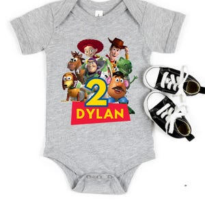 May include: A gray baby onesie featuring a colorful graphic of Toy Story characters, the number 2, and the name "Dylan" in red and yellow. Two black and white canvas sneakers with white laces are next to the onesie.