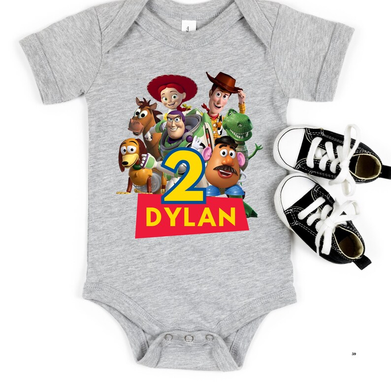 May include: A gray baby onesie featuring a colorful graphic of Toy Story characters, the number 2, and the name "Dylan" in red and yellow. Two black and white canvas sneakers with white laces are next to the onesie.