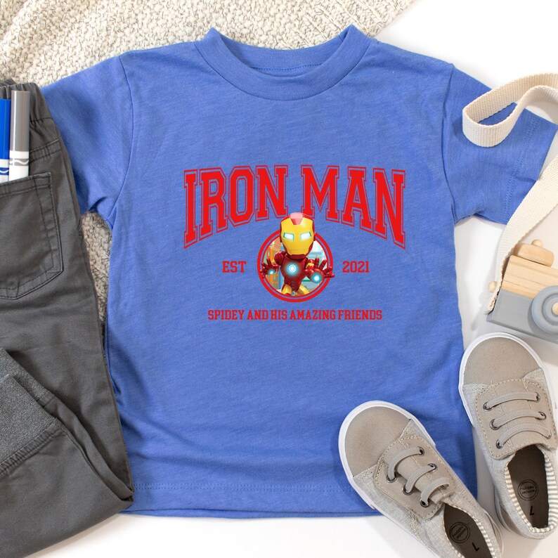 Pu&ograve; includere: Una maglietta blu con la scritta rossa "IRON MAN" e una grafica di Iron Man. La maglietta include anche la scritta "EST 2021" e "SPIDEY AND HIS AMAZING FRIENDS". Sono visibili anche pantaloni grigi e scarpe da ginnastica grigie.