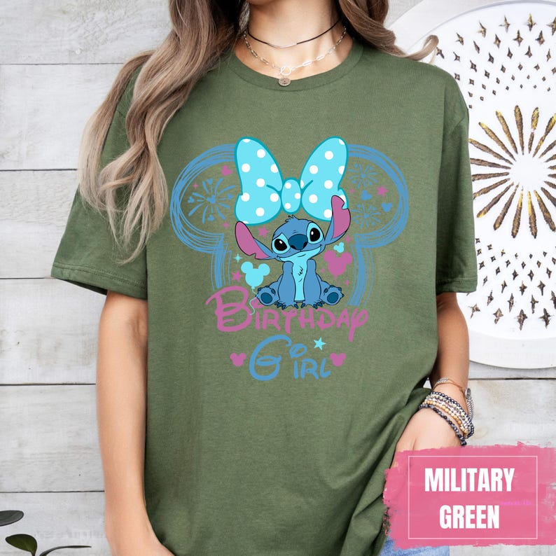 May include: Olive green t-shirt featuring a blue cartoon character with a polka dot bow, surrounded by a blue Mickey Mouse outline. The text "Birthday Girl" is in pink and blue. The bottom right corner has a pink rectangle with the words "MILITARY GREEN".