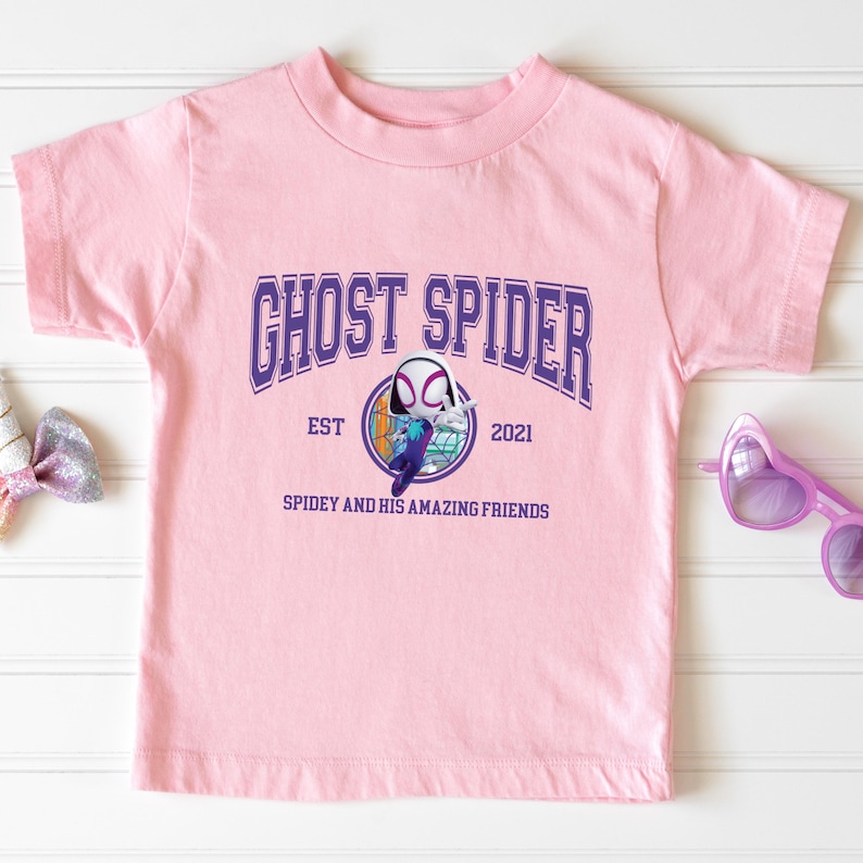 Pu&ograve; includere: T-shirt rosa chiaro con la scritta "GHOST SPIDER" in lettere viola. Sotto, una grafica di Ghost Spider e la scritta "SPIDEY AND HIS AMAZING FRIENDS". La maglietta ha girocollo e maniche corte.