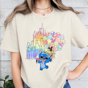 Puede incluir: Camiseta beige con un gr&aacute;fico colorido de Stitch de Lilo & Stitch sosteniendo un helado y globos en forma de coraz&oacute;n. Un castillo de colores arco&iacute;ris se encuentra en el fondo.