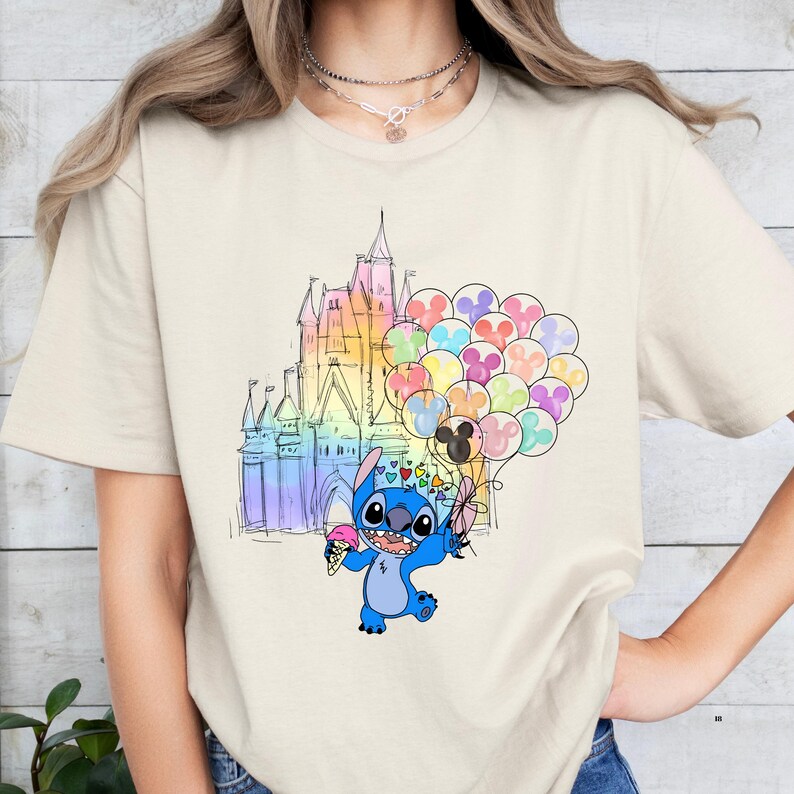 Puede incluir: Camiseta beige con un gr&aacute;fico colorido de Stitch de Lilo & Stitch sosteniendo un helado y globos en forma de coraz&oacute;n. Un castillo de colores arco&iacute;ris se encuentra en el fondo.
