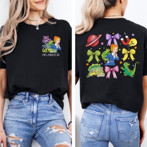 May include: Black t-shirt featuring a cartoon image of a woman in a blue dress with a green dinosaur and the text "TAKE CHANCES, MISTAKES, GET MESSY, MRS. ANDERSON" on the front. The back of the shirt has a colorful design with a planet, bows, and a school bus.