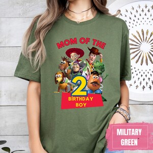 May include: Military green t-shirt featuring a graphic of Toy Story characters with the text "MOM OF THE" above the number "2" and "BIRTHDAY BOY" on a red rectangle. The shirt is a solid color.