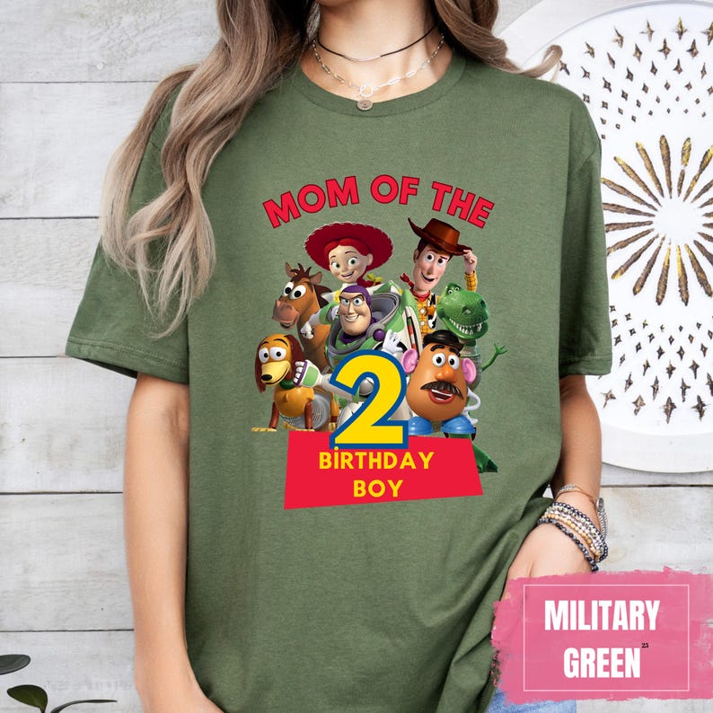 May include: Military green t-shirt featuring a graphic of Toy Story characters with the text "MOM OF THE" above the number "2" and "BIRTHDAY BOY" on a red rectangle. The shirt is a solid color.